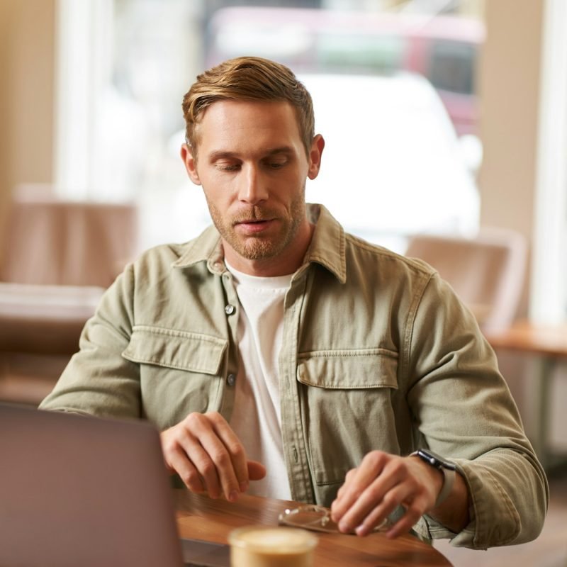 Image of young man, concentrated on his task, sitting with laptop in cafe, putting on glasses